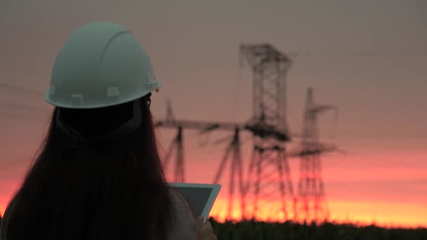 A woman power engineer in white helmet inspects power line using data from electrical sensors on a tablet. High voltage electrical lines at sunset. Distribution and supply of electricity. clean energy - Footage, Video