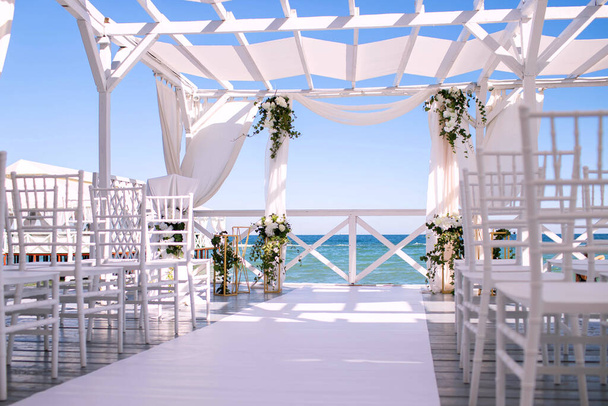 Весільна арка на тлі моря, свіжі квіти на декорі з білого дерева, красивий сонячний день для весільної церемонії, стільці для гостей, що чекають нареченого і нареченого
 - Фото, зображення