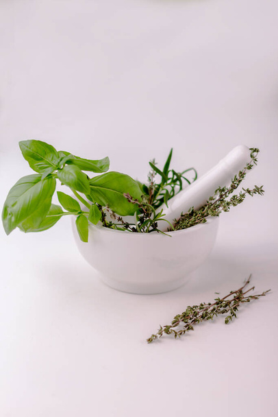 Green basil, thyme, oregano in white ceramic mortar and pestle om white table. Alternative herbal medicine concept. Vertical photo. Isolated on white. Selective focus. - Фото, изображение