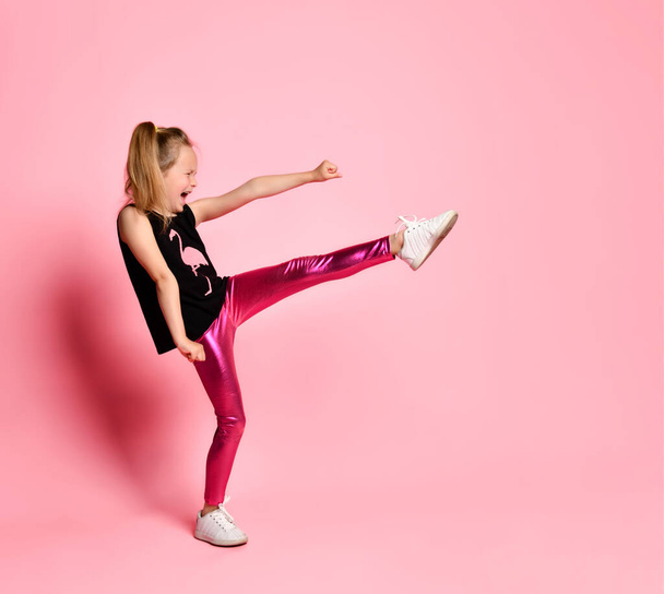 Capricious little girl in black t-shirt and glossy leggings screaming loudly, kicking and punching. Full length shot isolated on pink, copy space - Foto, Imagem