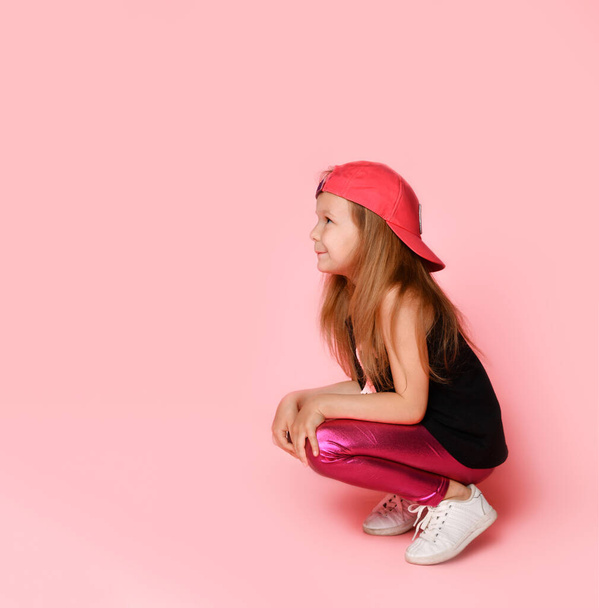Charming active preschooler in trendy trendy dress, playful and smiling, squatting, looking to the side, on a pink background - 写真・画像
