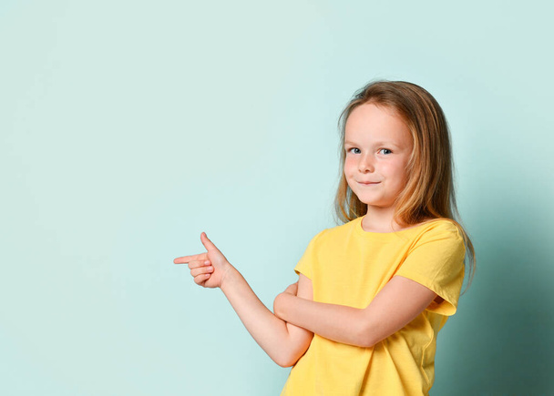 Little female in yellow t-shirt. She is smiling and pointing to the side by forefinger while posing against turquoise studio background. Close up - Foto, Imagen