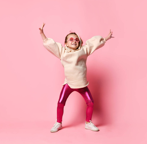 Blonde cheerful girl posing in a stylish outfit of leggings and hoodies. Happy teenage boy posing in studio in pink glasses. Bright positive colors. - Foto, Imagem