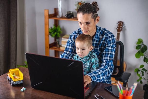 若い現代人の父はノートパソコンで働いていて、彼の小さな息子は彼の膝の上に座っています。家庭からの仕事とリモートワークの概念 - 写真・画像