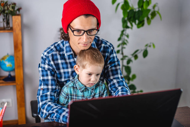 Jovem pai homem moderno está trabalhando em um laptop, e seu filho está sentado em seu colo. Conceito de trabalho familiar e remoto em casa - Foto, Imagem