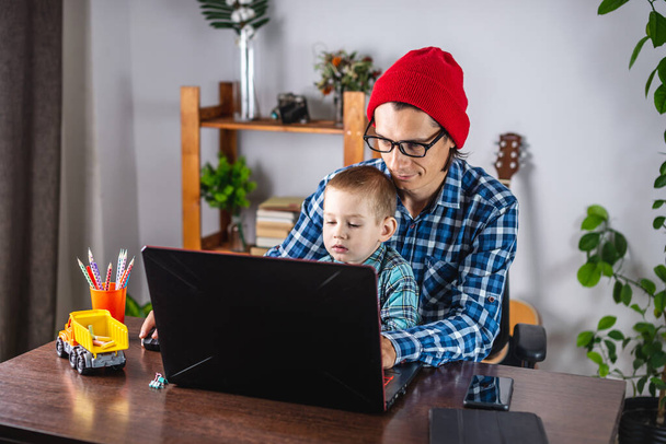 Le jeune homme moderne père travaille sur un ordinateur portable, et son petit fils est assis sur ses genoux. Concept de famille et de travail à distance à domicile - Photo, image
