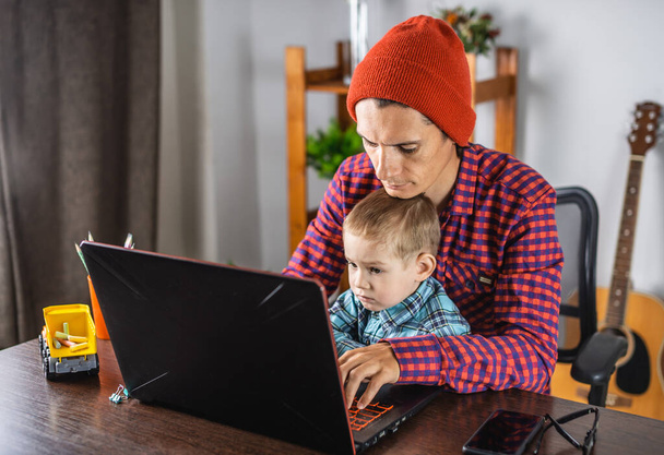 Il padre giovane uomo moderno sta lavorando su un computer portatile, e il suo piccolo figlio è seduto sulle sue ginocchia. Concetto di famiglia e lavoro a distanza da casa - Foto, immagini