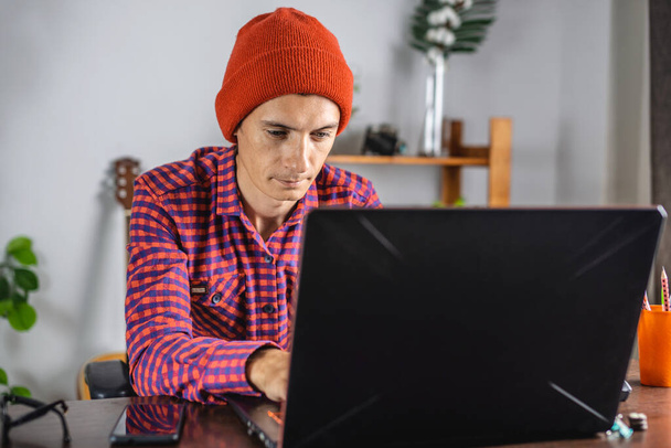 Сучасний молодий чоловік у червоній перевіреній сорочці і капелюсі працює на ноутбуці. Концепція дистанційного зв'язку та віддаленої роботи
. - Фото, зображення