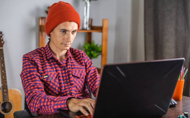 Сучасний молодий чоловік у червоній перевіреній сорочці і капелюсі працює на ноутбуці. Концепція дистанційного зв'язку та віддаленої роботи
. - Фото, зображення