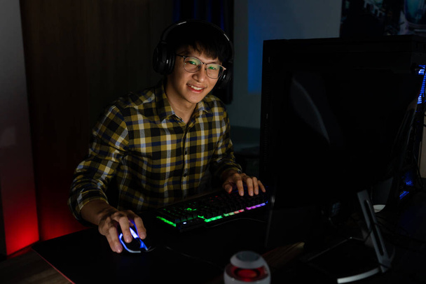 Knappe opgewonden aziatische gamer man in hoofdtelefoon genieten en verheugen tijdens het spelen van videospelletjes op de computer in gezellige kamer is verlicht met warm en neon licht, gaming en technologie e-sport concept. - Foto, afbeelding