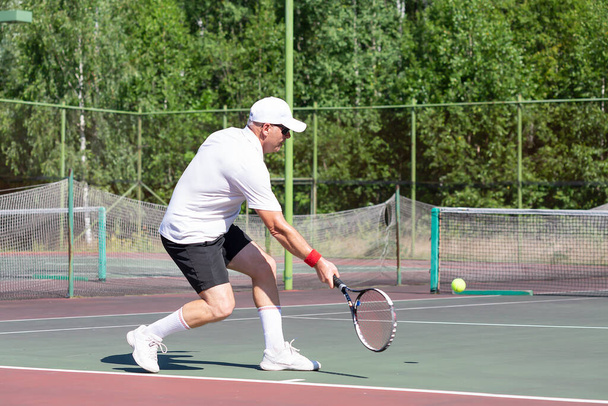 An elderly man in good physical shape plays tennis on an outdoor court on a sunny day - Fotoğraf, Görsel