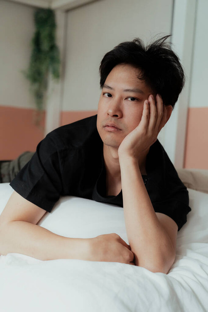 Ázsiai meleg fekete pólóban feküdjön a fehér ágyra egy párnával a mellkasa alatt nézzen a kamerába unatkozó látással.. - Fotó, kép