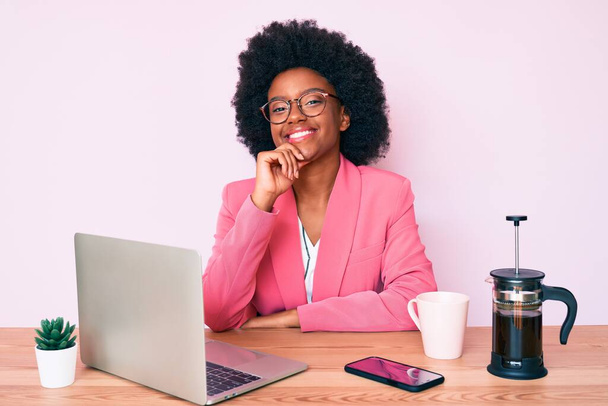 Joven mujer afroamericana que trabaja en el escritorio usando computadora portátil sonriendo mirando confiado a la cámara con los brazos cruzados y la mano en la barbilla. pensar positivamente.  - Foto, Imagen