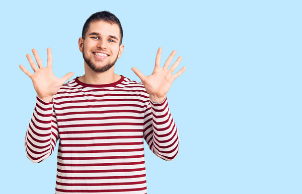 Νεαρός όμορφος άντρας με ριγέ πουλόβερ που δείχνει και δείχνει προς τα πάνω με τα δάχτυλα νούμερο δέκα ενώ χαμογελά σίγουρος και χαρούμενος.  - Φωτογραφία, εικόνα
