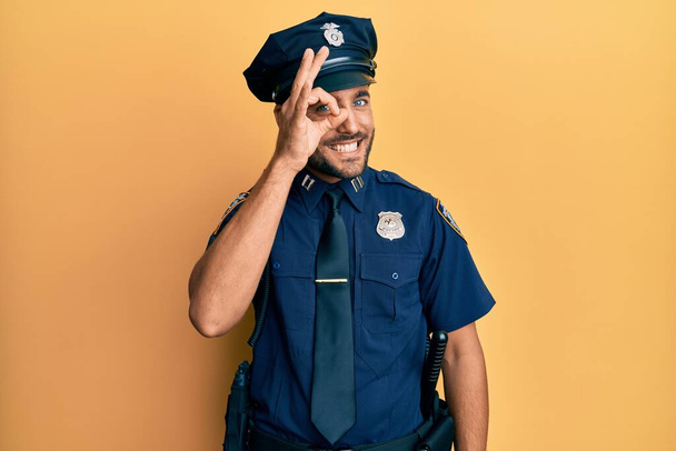 Handsome hispanic man wearing police uniform doing ok gesture with hand smiling, eye looking through fingers with happy face.  - Foto, Imagem