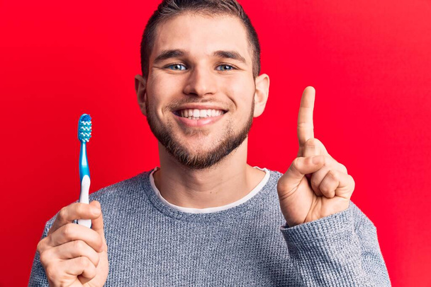 Junger gutaussehender Mann mit Zahnbürste, der mit einer Idee oder Frage lächelt und mit glücklichem Gesicht den Finger zeigt, Nummer eins  - Foto, Bild