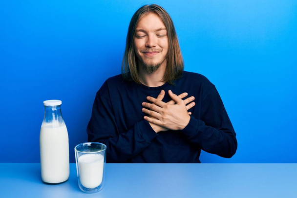 Красивий білий чоловік з довгим волоссям тримає склянку молока, посміхаючись руками на грудях, очі закриті вдячним жестом на обличчі. концепція здоров'я
.  - Фото, зображення