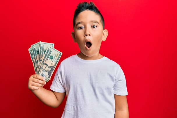 Kleiner hispanischer Junge, der 20-Dollar-Banknoten in der Hand hält, erschrocken und erstaunt mit offenem Mund zur Überraschung, ungläubiges Gesicht  - Foto, Bild