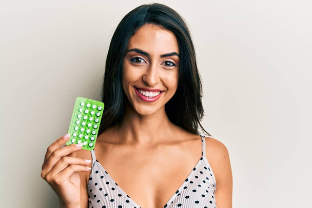 Beautiful hispanic woman holding birth control pills looking positive and happy standing and smiling with a confident smile showing teeth  - Foto, Imagem
