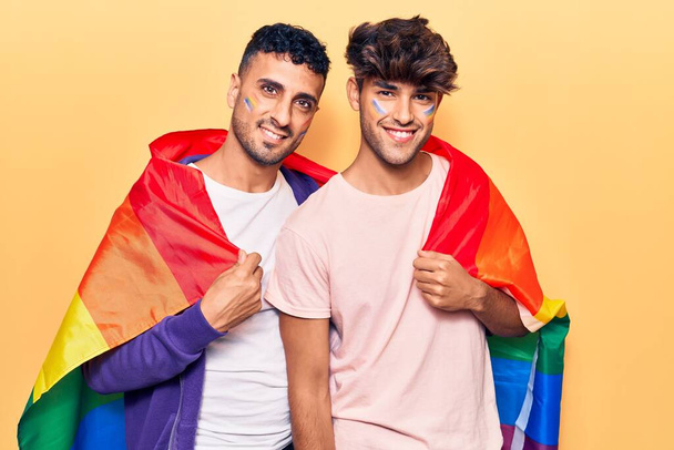 Mladý gayové pár nosí duhu lgbtq vlajka vypadá pozitivní a šťastný stojící a usmívá se s sebevědomým úsměvem ukazující zuby  - Fotografie, Obrázek