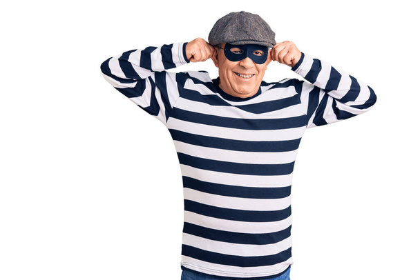 Homem bonito sênior vestindo máscara de ladrão e t-shirt sorrindo puxando orelhas com os dedos, gesto engraçado. problema de audição  - Foto, Imagem