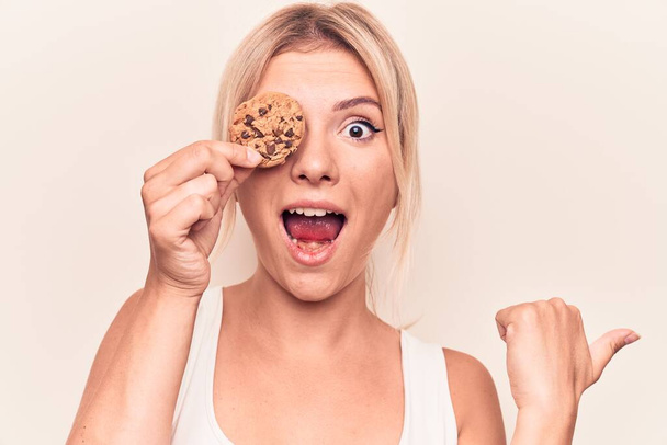 Mladá krásná blondýnka žena drží sladkou čokoládovou sušenku přes oko ukazující palec nahoru na stranu s úsměvem šťastný s otevřenými ústy - Fotografie, Obrázek