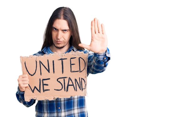 Молодой взрослый человек с длинными волосами держась вместе мы стоим баннер с открытой рукой делать стоп-знак с серьезным и уверенным выражением, защитный жест  - Фото, изображение