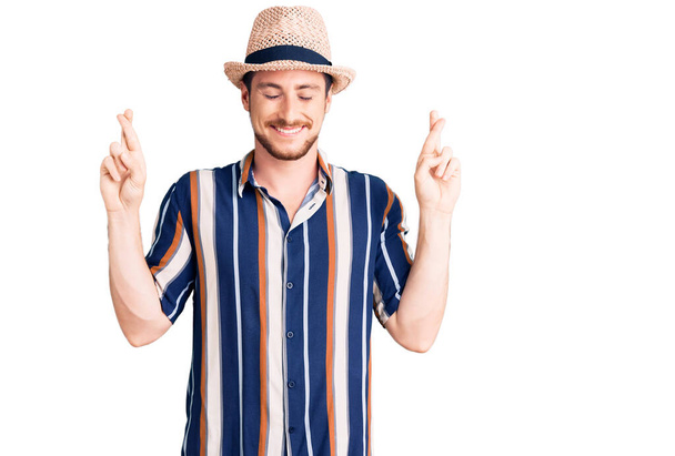 Young handsome caucasian man wearing summer hat gesturing finger crossed smiling with hope and eyes closed. luck and superstitious concept.  - Fotoğraf, Görsel