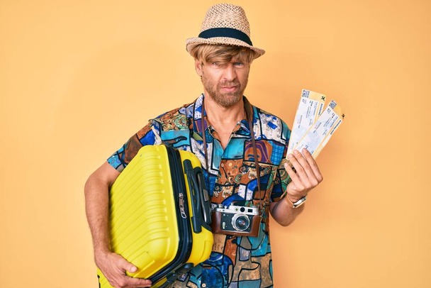 Junger blonder Mann in Sommerkleidung mit Handtasche und Bordkarte, skeptisch und nervös, die Stirn runzelnd wegen des Problems. Negative Person.  - Foto, Bild