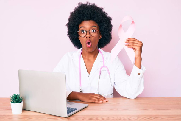 Genç Afro-Amerikan kadın doktor üniforması giyiyor elinde pembe kanser kurdelesi var korkmuş ve ağzı açık şaşkınlık içinde, inançsız bir yüz.  - Fotoğraf, Görsel