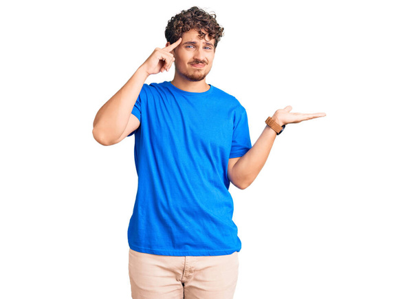 Young handsome man with curly hair wearing casual clothes confused and annoyed with open palm showing copy space and pointing finger to forehead. think about it.  - Photo, Image