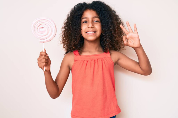 Enfant afro-américain aux cheveux bouclés tenant la sucette faisant signe ok avec les doigts, geste amical souriant excellent symbole  - Photo, image