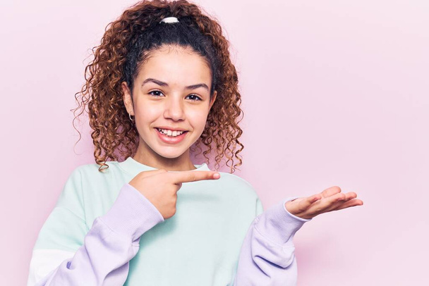Beautiful kid girl with curly hair wearing casual clothes amazed and smiling to the camera while presenting with hand and pointing with finger.  - Photo, Image