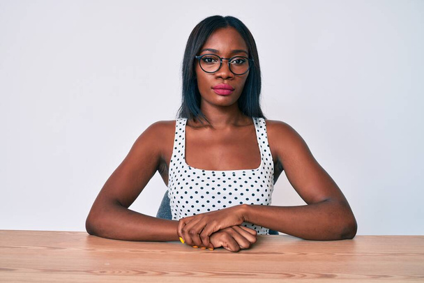 Nuori afrikkalainen amerikkalainen nainen päällään rento vaatteita ja lasit istuu pöydällä vakava ilme kasvoilla. yksinkertainen ja luonnollinen kameraan katsominen.  - Valokuva, kuva