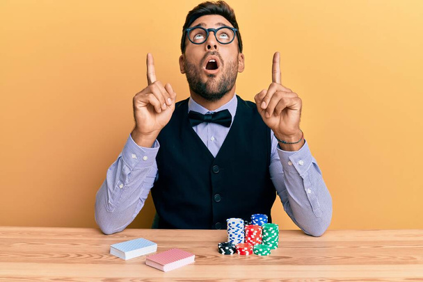 Przystojny Latynos krupier siedzi na stole z żetonów i kart pokerowych zdumiony i zaskoczony patrząc w górę i wskazując palcami i podniesionymi ramionami.  - Zdjęcie, obraz