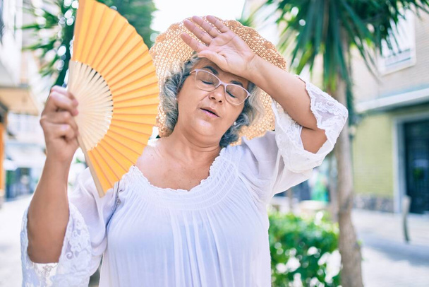 Kobieta w średnim wieku z siwymi włosami używająca ręcznego wentylatora w bardzo gorący dzień fali upałów - Zdjęcie, obraz