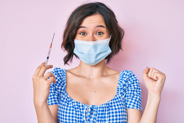 Young beautiful woman wearing medical mask holding syringe screaming proud, celebrating victory and success very excited with raised arms  - Photo, Image