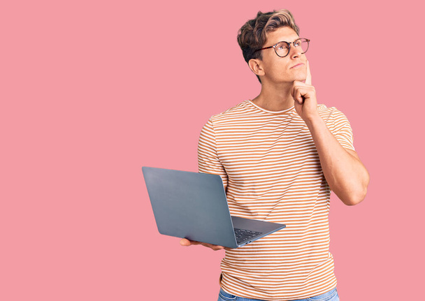 Fiatal jóképű férfi szemüvegben tartja laptop komoly arc gondolkodás kérdés kézzel az állán, gondolkodó zavaros ötlet  - Fotó, kép