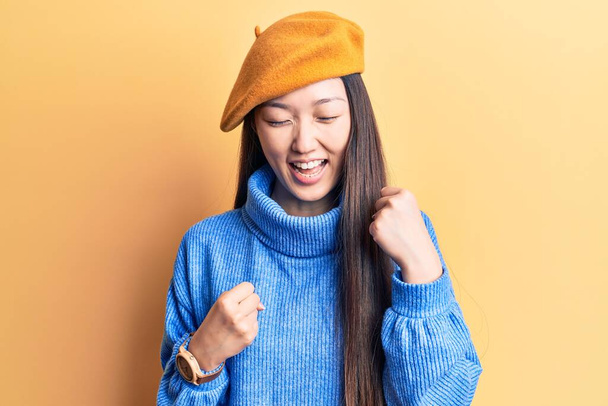 Jovem mulher chinesa bonita vestindo camisola gola alta e boina francesa comemorando surpreso e surpreso com o sucesso com os braços levantados e os olhos fechados  - Foto, Imagem