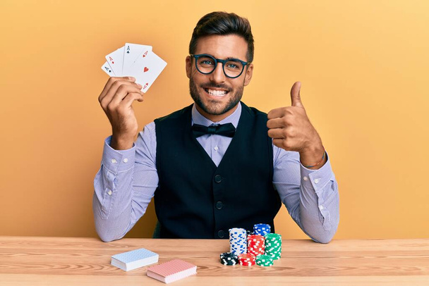 Hombre guapo croupier hispano sentado en la mesa con fichas de póquer y tarjetas sonriendo feliz y positivo, pulgar hacia arriba haciendo excelente y signo de aprobación  - Foto, Imagen