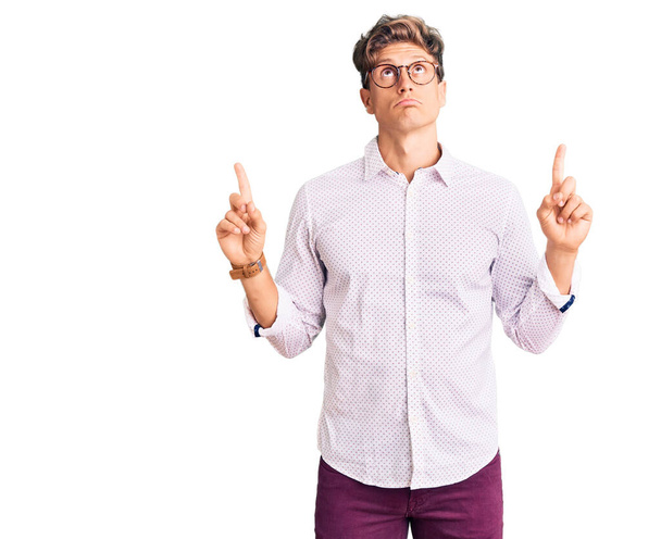 Homem bonito jovem vestindo roupas de negócios e óculos apontando para cima olhando triste e chateado, indicando direção com os dedos, infeliz e deprimido.  - Foto, Imagem