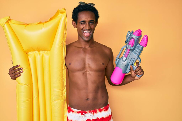 African handsome man wearing swimsuit and holding summer float winking looking at the camera with sexy expression, cheerful and happy face.  - Фото, зображення