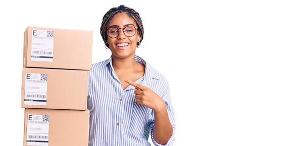 Молода американка з плечима, що тримає коробки для доставки, посміхається, показуючи пальцем руки.  - Фото, зображення