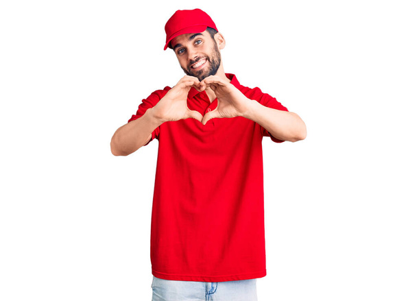Jeune homme beau avec barbe portant uniforme de livraison souriant dans l'amour faisant forme de symbole de coeur avec les mains. concept romantique.  - Photo, image