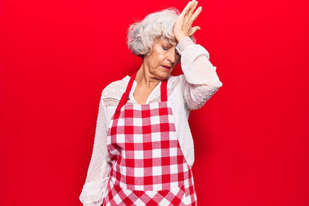 Oudere grijsharige vrouw die een schort draagt verrast met hand op hoofd voor fout, onthoud fout. vergeten, slecht geheugen concept.  - Foto, afbeelding