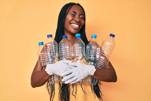 Африканская американка с косичками держа утилизирующие пластиковые бутылки подмигивая глядя на камеру с сексуальным выражением лица, веселым и счастливым лицом.  - Фото, изображение