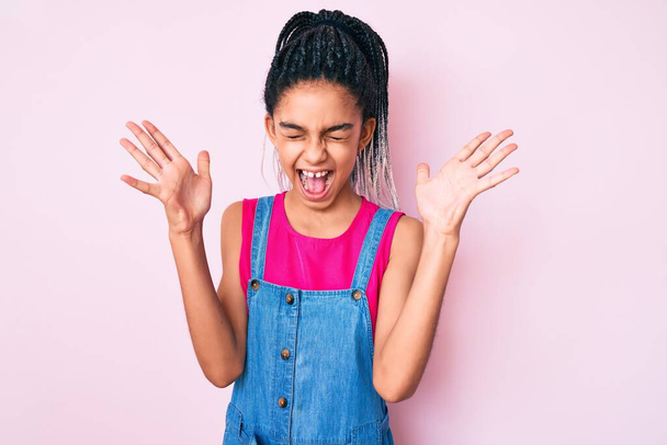 Młody Afroamerykanin dziewczyna dziecko z warkoczami noszenie ubrań na różowym tle świętując szalony i szalony dla sukcesu z ramionami podniesione i zamknięte oczy krzycząc podekscytowany. koncepcja zwycięzcy  - Zdjęcie, obraz