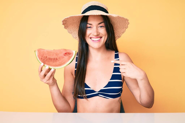 Jeune belle femme caucasienne portant un bikini et un chapeau tenant une tranche de pastèque pointant du doigt vers un sourire heureux et fier  - Photo, image