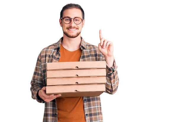 Молодой красивый белый мужчина с коробкой пиццы, удивленный идеей или вопросом, указывая пальцем со счастливым лицом, номер один  - Фото, изображение