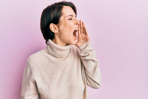 Young brunette woman with short hair wearing casual winter sweater shouting and screaming loud to side with hand on mouth. communication concept.  - Photo, Image
